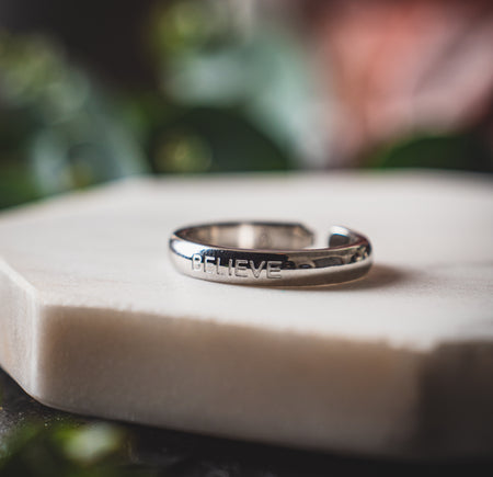 Shiny stainless steel ring with the word 'BELIEVE' engraved, placed on a soft-textured white surface with a blurred background of greenery.