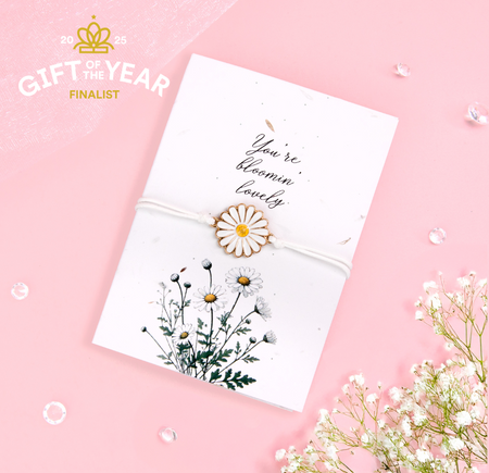 A delicate daisy bracelet featuring a white and yellow flower charm, tied with a soft white cord, displayed on a seeded card with the text