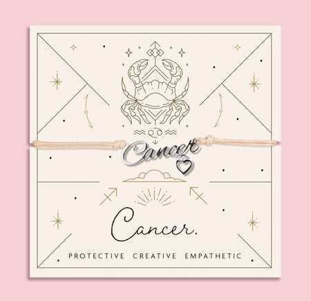 Adjustable beige cord bracelet with a silver 'Cancer' charm and small heart pendant, displayed on a zodiac-themed card featuring a crab illustration and the traits