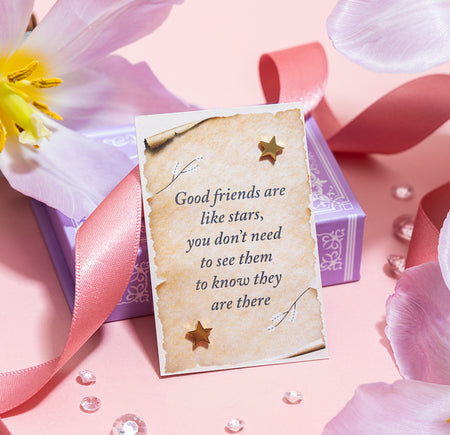 A pair of simple gold star-shaped earrings presented in an open purple gift box, which features an inspiring quote on a parchment-style card saying, 