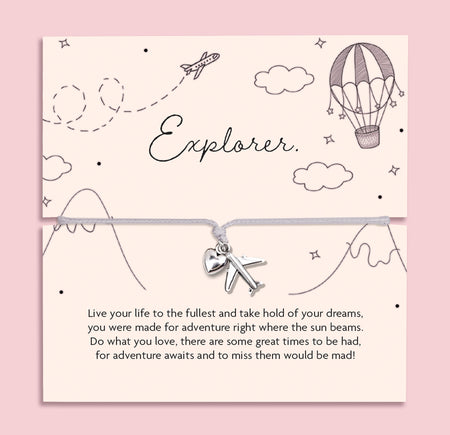 Flat lay of a silver charm bracelet featuring a heart and airplane charm, displayed on a pastel card with hand-drawn travel illustrations and the word