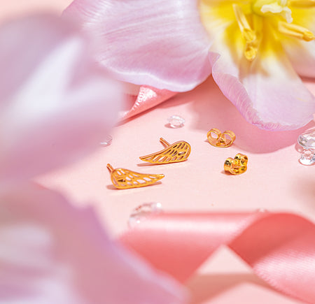 Close-up image of tiny gold feather-shaped stud earrings with butterfly backs, styled on a soft pink background with floral petals and satin ribbon, part of the Letterbox Love earring collection.