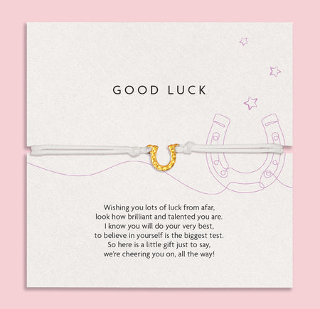 Charming 'GOOD LUCK' card on a soft pink background, adorned with encouraging words and a horseshoe illustration, accompanied by a slender white bracelet with a golden horseshoe charm.