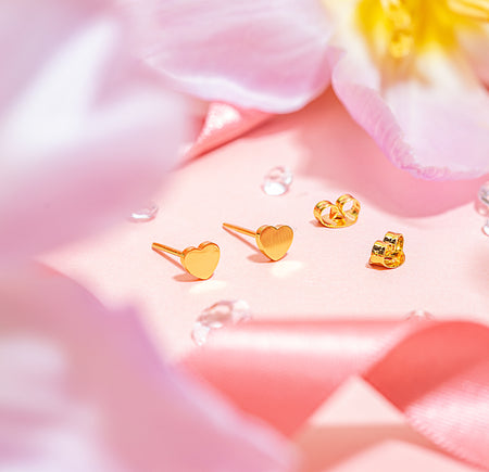 Close-up image of tiny gold heart stud earrings with matching butterfly backs, displayed on a pink background with floral and ribbon accents, from the Letterbox Love earring collection.