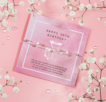 A 'Happy 30th Birthday' bracelet with silver star charms, displayed on a card with a celebratory message, amidst a backdrop of pink hues and white baby's breath flowers, symbolizing the joy and milestone of turning thirty.