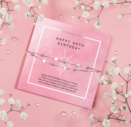 A celebratory 60th birthday bracelet by Letterbox Love, displayed on a soft pink background with glistening white flowers. The bracelet features a silver chain interspersed with shimmering stars and beads, conveying elegance and joy. It lies atop a matching pink birthday card that reads 