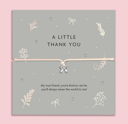 A flat-lay image of a 'A Little Thank You' bracelet gift card. The card is grey with delicate cream botanical and heart illustrations, featuring a thin cream cord bracelet with a small silver bow charm in the center. Below the bracelet, there's a sentimental message: 'My true friend, you're kind as can be, you'll always mean the world to me!' The background is a soft pink.