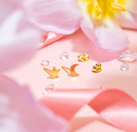 Gold-plated stainless steel star and crescent moon stud earrings with butterfly backs, styled on a soft pink background with floral accents and ribbon.