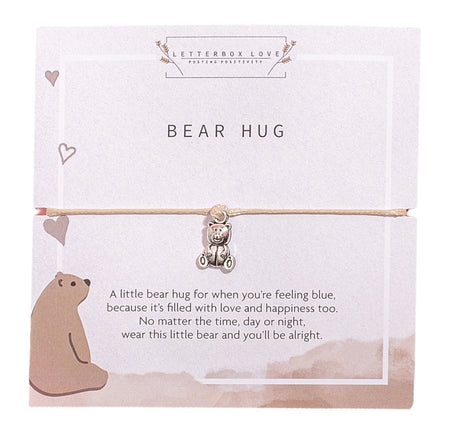 A light cream and beige card with the title 'Bear Hug' at the top. It features a silver teddy bear charm attached to a beige cord bracelet. The card has a cute illustration of a bear sitting in the bottom left corner with small heart accents. Below the bracelet, a comforting message reads: 'A little bear hug for when you’re feeling blue, because it’s filled with love and happiness too. No matter the time, day or night, wear this little bear and you’ll be alright.