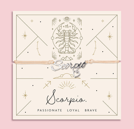Scorpio zodiac bracelet with silver calligraphy-style “Scorpio” charm and dangling heart on a beige cord, displayed on a celestial cream and gold card with traits: Passionate, Loyal, Brave.