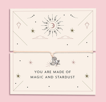 Flat lay image of a delicate cord bracelet with silver moon and star charms, displayed on a celestial-themed card that reads “You are made of magic and stardust.” The card features star and sun illustrations, set against a soft cream background with a pink border.