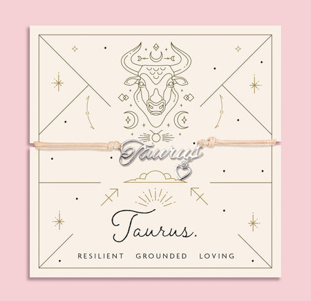 Adjustable peach cord bracelet with a silver 'Taurus' charm and heart pendant, displayed on a zodiac-themed backing card with a bull illustration and traits reading