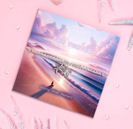 A silver-plated bracelet with a delicate turtle and starfish charm, displayed on a serene beach-themed backing card featuring a pink and purple sunrise over the ocean. The text reads