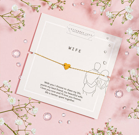 A dainty gold bracelet with a small, solid heart charm presented on a white card with the word 'WIFE' in bold, centered text. Below the bracelet, a loving message to a wife is printed, expressing lifelong commitment and affection.
