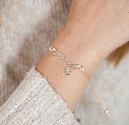 A woman wearing a delicate bracelet with the word “Pisces” and a small heart charm, a meaningful zodiac bracelet gift for her.