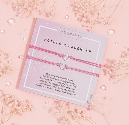 Pair of pink corded bracelets with heart-shaped charms presented on a lavender 'MOTHER & DAUGHTER' card, with a touching message about love and togetherness, adorned with baby's breath flowers and crystal accents on a pastel pink backdrop.
