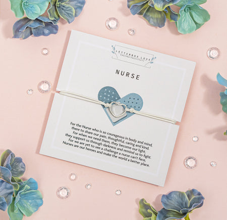 White bracelet with a silver heart-shaped charm showcased on a white 'NURSE' appreciation card with a heartfelt message about their courage and care, surrounded by blue-green flowers and crystal accents on a soft pink background