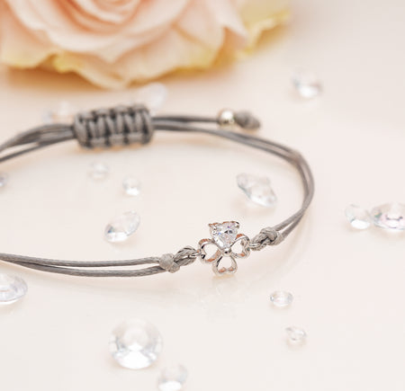 The image shows a delicate bracelet placed on a soft, light-colored surface with a blurred pink rose in the background. The bracelet features a silver four-leaf clover charm with a sparkling heart-shaped gem at its center, attached to a grey cord. The cord is tied in an adjustable knot design for easy resizing. Scattered around the bracelet are small, clear decorative gems, enhancing the elegant and charming aesthetic of the piece.