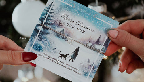 Close-up of a "Merry Christmas Dog Mum" bracelet card held in front of a Christmas tree, featuring a paw charm and festive winter scene.