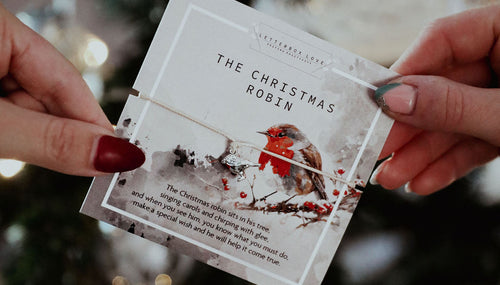 Two women's hands holding a "Christmas Robin" gift card with a string friendship bracelet with a cute robin charm