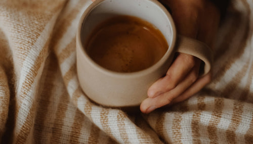 Close-up of a person’s hands holding a warm mug of coffee while wrapped in a soft, striped blanket, evoking feelings of coziness and relaxation during a chilly autumn day.