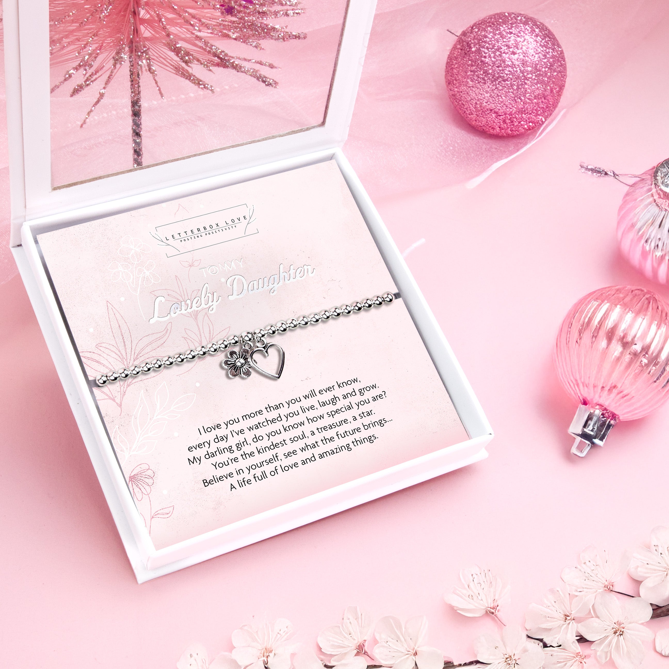 A pink, festive set-up with a Letterbox Love Gift box featuring a silver beaded bracelet. The bracelet is silver-plated with a heart and small flower charm. It has a sentimental message titled 