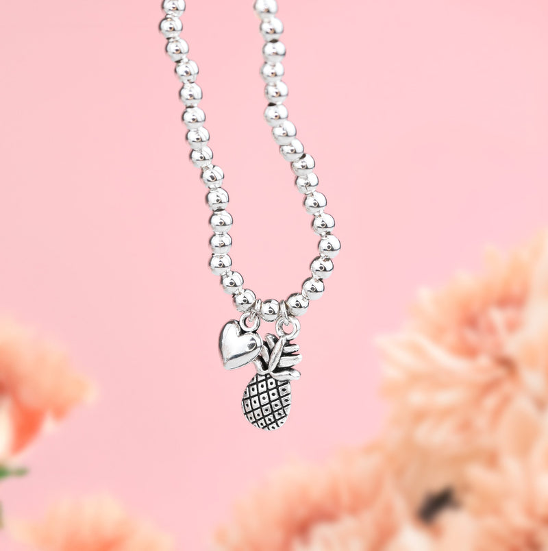 Pink background and blurred flowers with a silver-plated beaded bracelet with pineapple and heart charms. A gift for someone going through IVF treatment.
