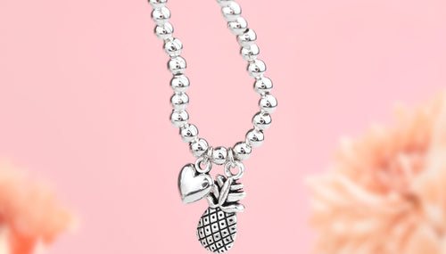Pink background and blurred flowers with a silver-plated beaded bracelet with pineapple and heart charms. A gift for someone going through IVF treatment.