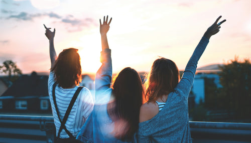 three women with friendship bracelets staring into the sunset with their arms in the air