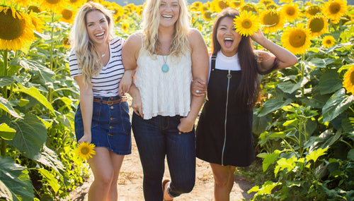 three women walking through a sunflower field with linked arms and laughing