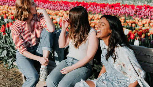 Three friends sitting on a bench in a vibrant tulip field, laughing and enjoying each other's company. They are dressed casually, showcasing a joyful and carefree moment.