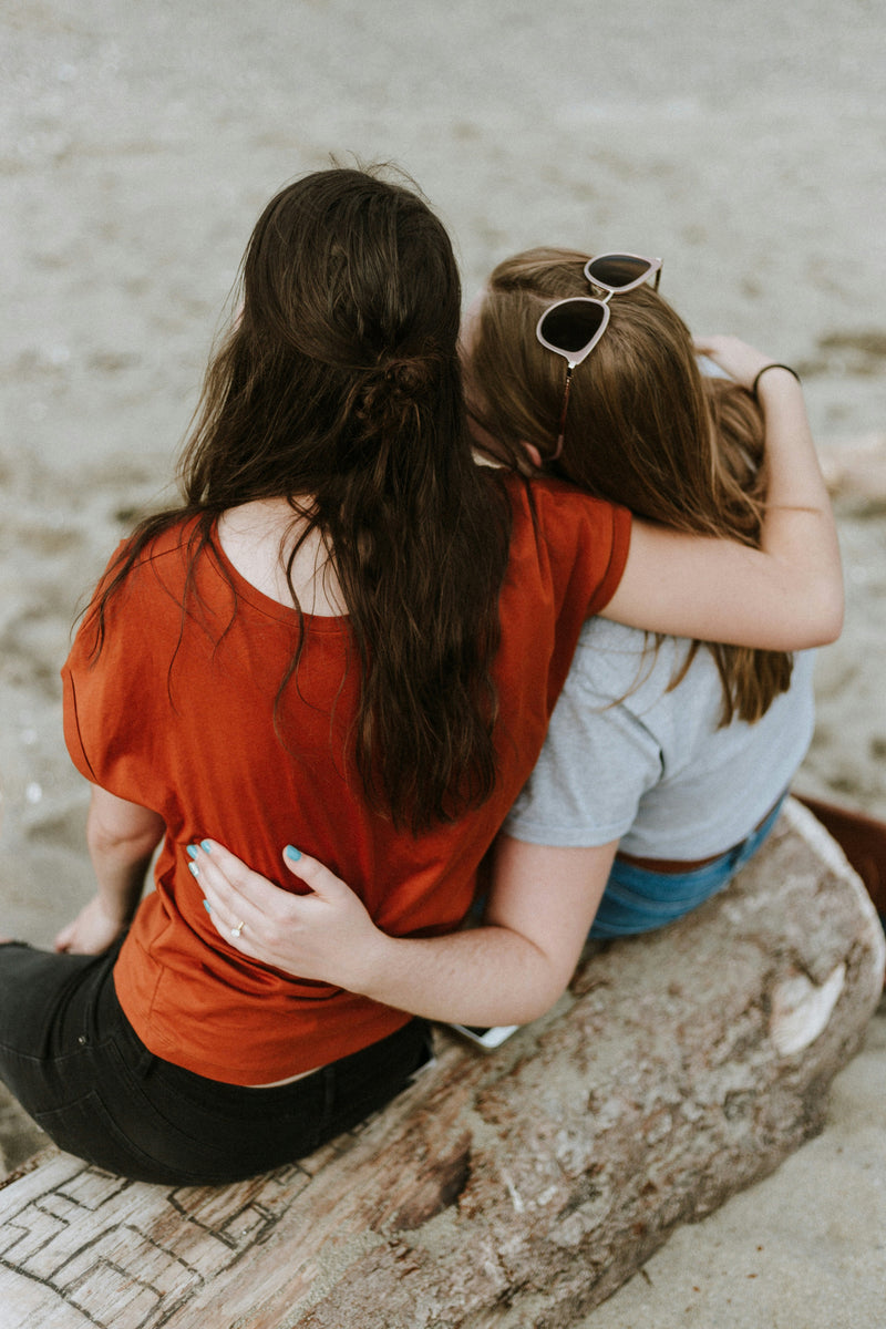 Two women embracing on a log on the beach. A woman with dark brown hair is wearing an orange top and a dark blonde haired woman has sunglasses on her head.