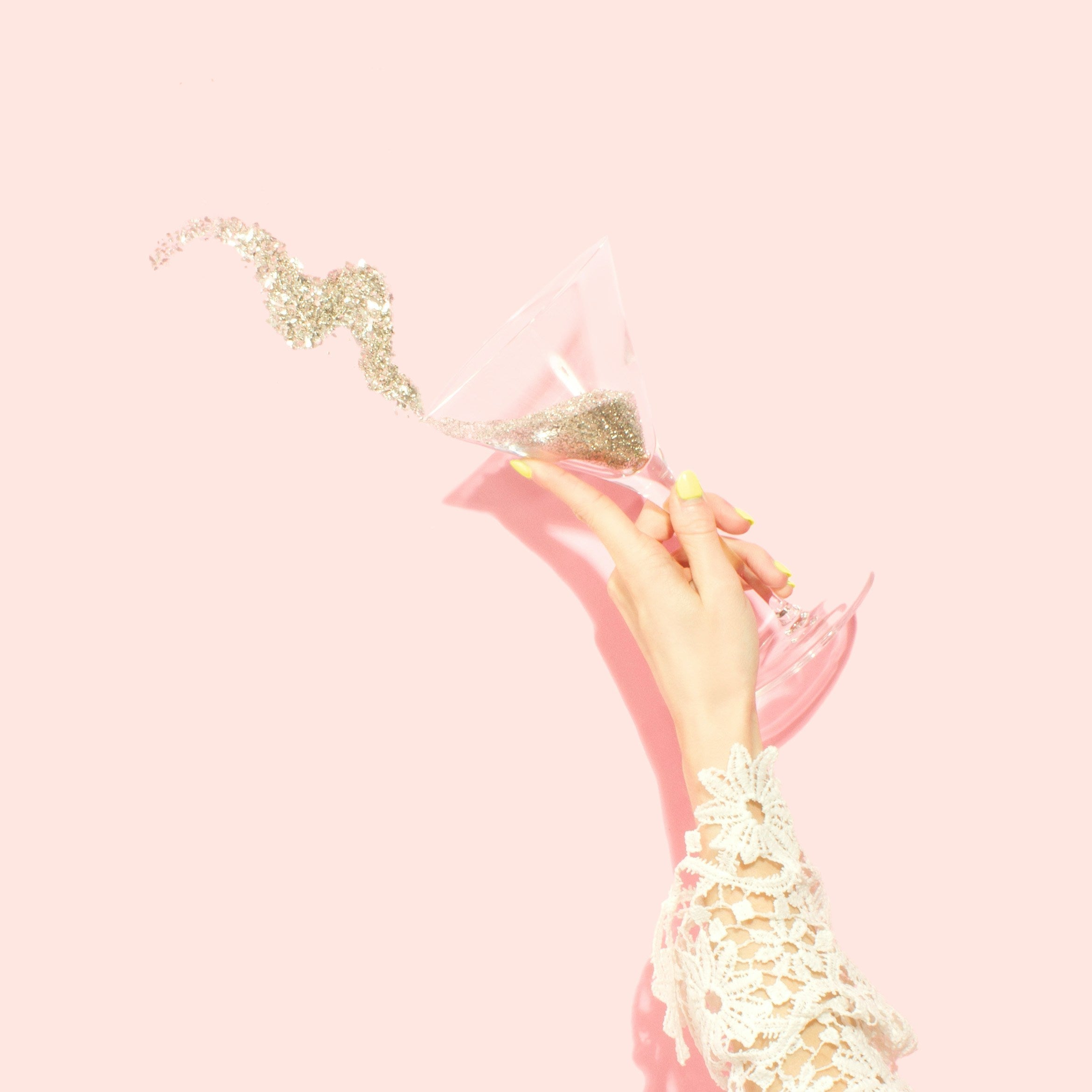 pale pink background with a woman's hand splashing gold sparkles from a martini glass