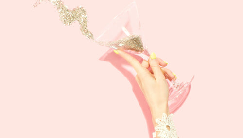 pale pink background with a woman's hand splashing gold sparkles from a martini glass