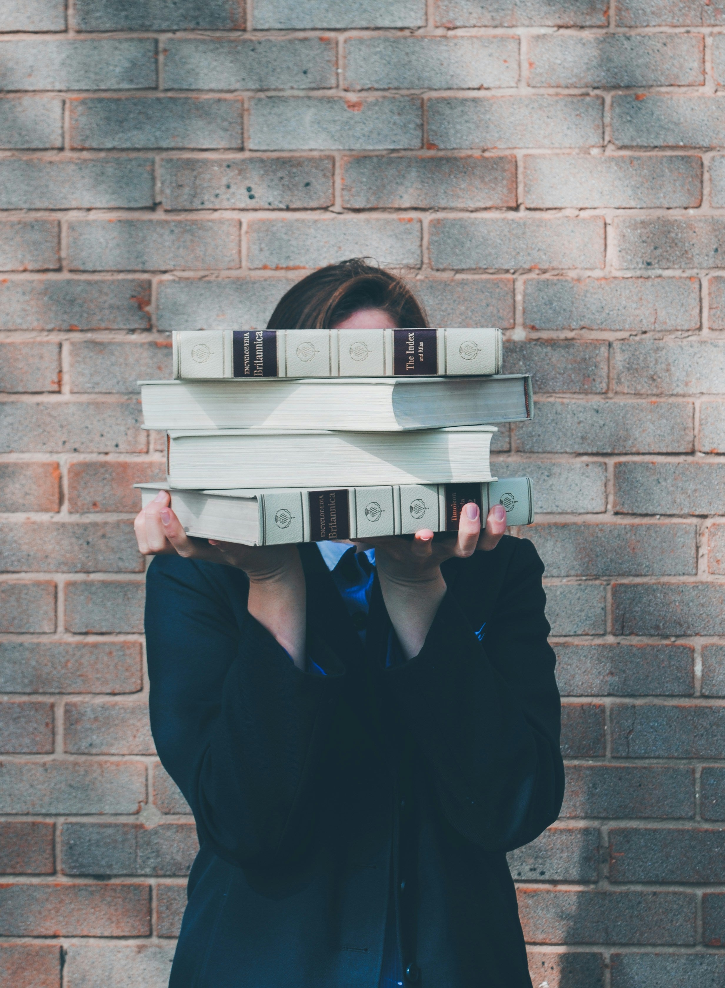 a girl stood in front of a grey brick wall holding academic books in front of her face. 