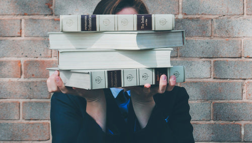 a girl stood in front of a grey brick wall holding academic books in front of her face. 
