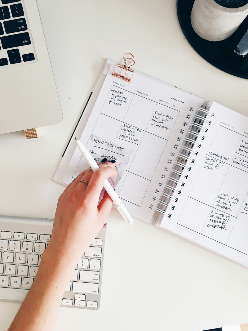 A woman at work with a white desk and keypad. Her planner is open with various tasks written and her hand and pen over the book.