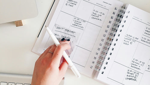 A woman at work with a white desk and keypad. Her planner is open with various tasks written and her hand and pen over the book.