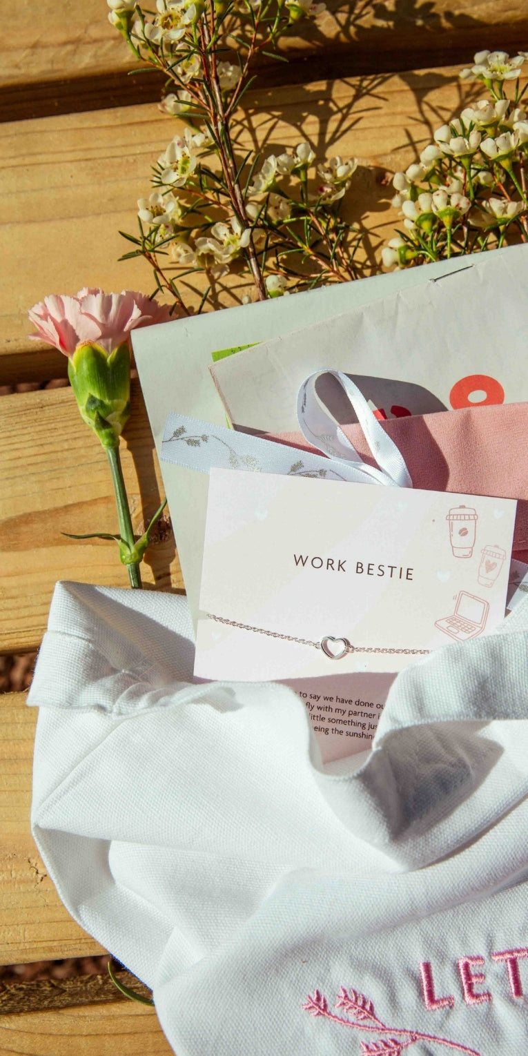 A wooden bench with a Letterbox Love tote bag on with a newspaper, velvet pouch and 'work bestie' bracelet falling out of the bag
