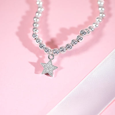 a pink background with a silver beaded star bracelet and white ribbon
