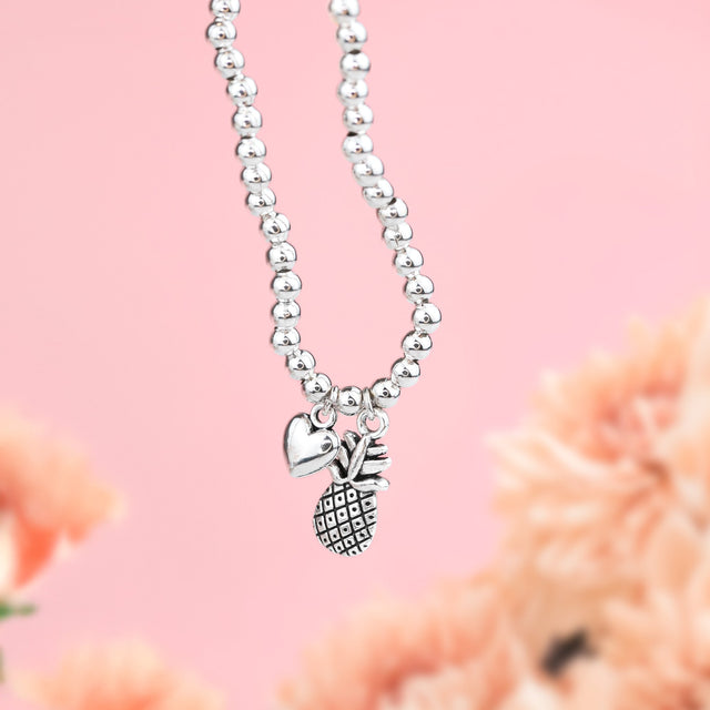 A pink background and blurred flowers with a silver-plated beaded bracelet with beautiful pineapple and heart charm. Gifts for those on an IVF Journey.