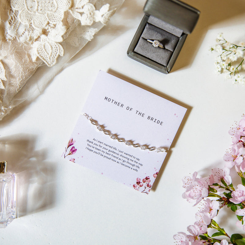 A white background with wedding dress and flowers, also a Miss Dior perfume bottle and wedding ring box. The Mother of the Bride bracelet gift is in the centre