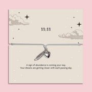 White adjustable 11:11 angel number bracelet with silver-tone feather and heart charms, displayed on a celestial sentiment card with a manifestation and abundance message.