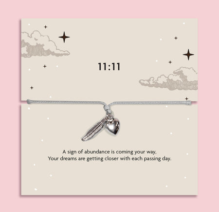 White adjustable 11:11 angel number bracelet with silver-tone feather and heart charms, displayed on a celestial sentiment card with a manifestation and abundance message.