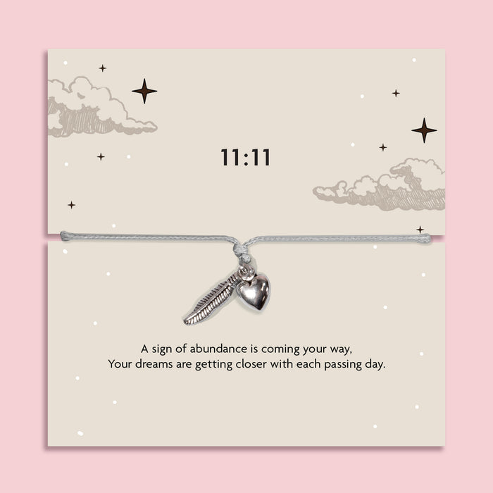 White adjustable 11:11 angel number bracelet with silver-tone feather and heart charms, displayed on a celestial sentiment card with a manifestation and abundance message.