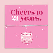 Pink 21st birthday gift card with the text “Cheers to 21 years” in bold pink and white lettering, featuring a silver and grey adjustable heart charm bracelet across the centre and an illustrated birthday cake underneath.