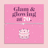 Pink 70th birthday gift card with the text “Glam & glowing at 70” in bold purple and white lettering, featuring a silver and grey adjustable heart charm bracelet across the centre and an illustrated birthday cake below.