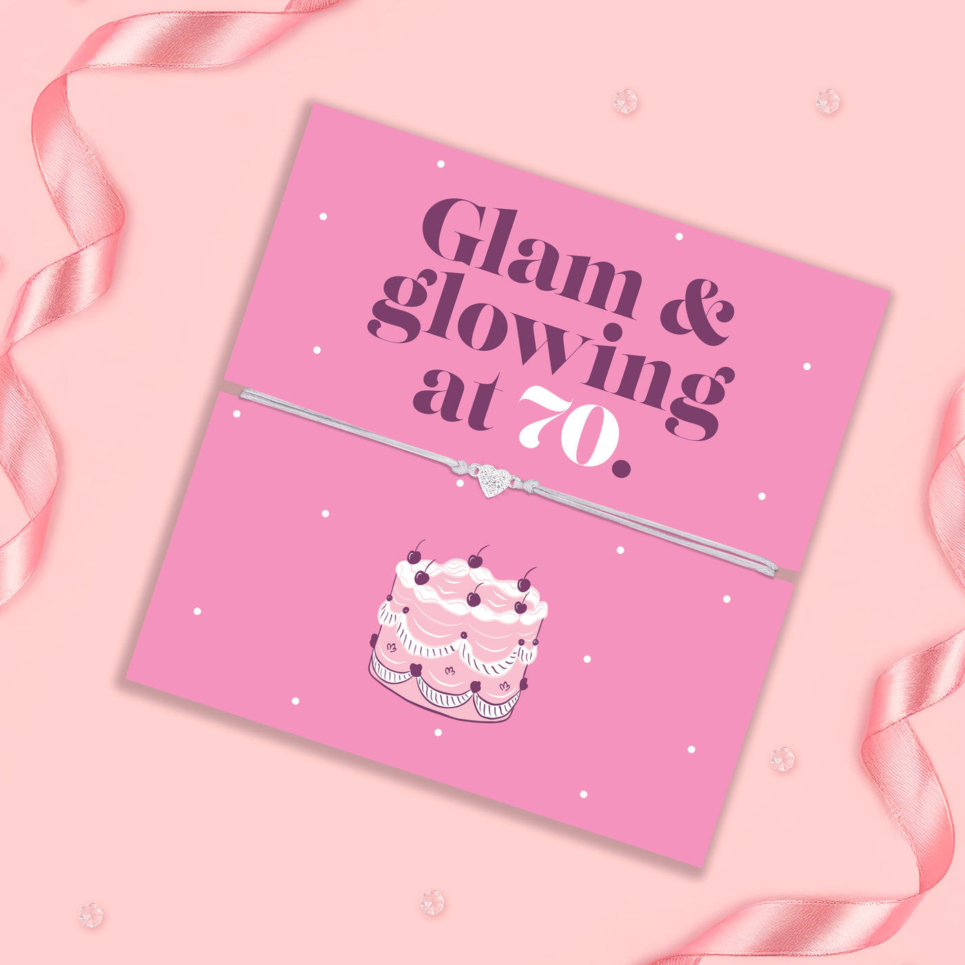 Pink birthday card with cake illustration and text 'Glam & glowing at 70' on a pink background with ribbons.