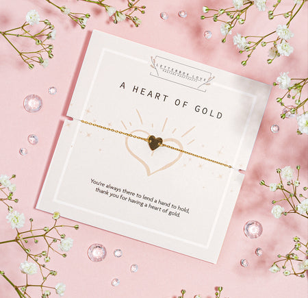 Gold heart charm bracelet presented on a 'Heart of Gold' card with an appreciative message, surrounded by delicate baby's breath on a soft pink backdrop with scattered crystals, conveying gratitude and affection.