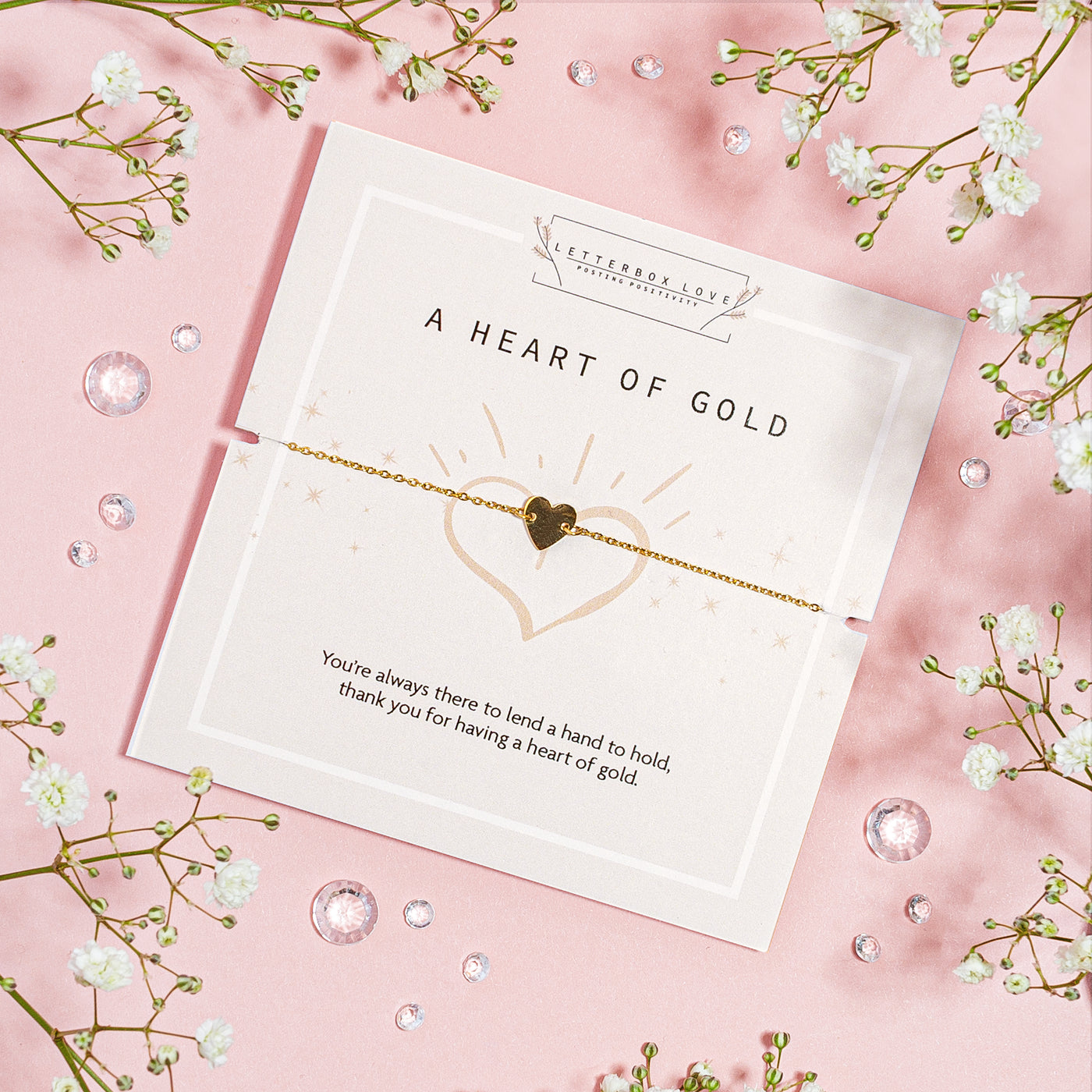 Gold heart charm bracelet presented on a 'Heart of Gold' card with an appreciative message, surrounded by delicate baby's breath on a soft pink backdrop with scattered crystals, conveying gratitude and affection.